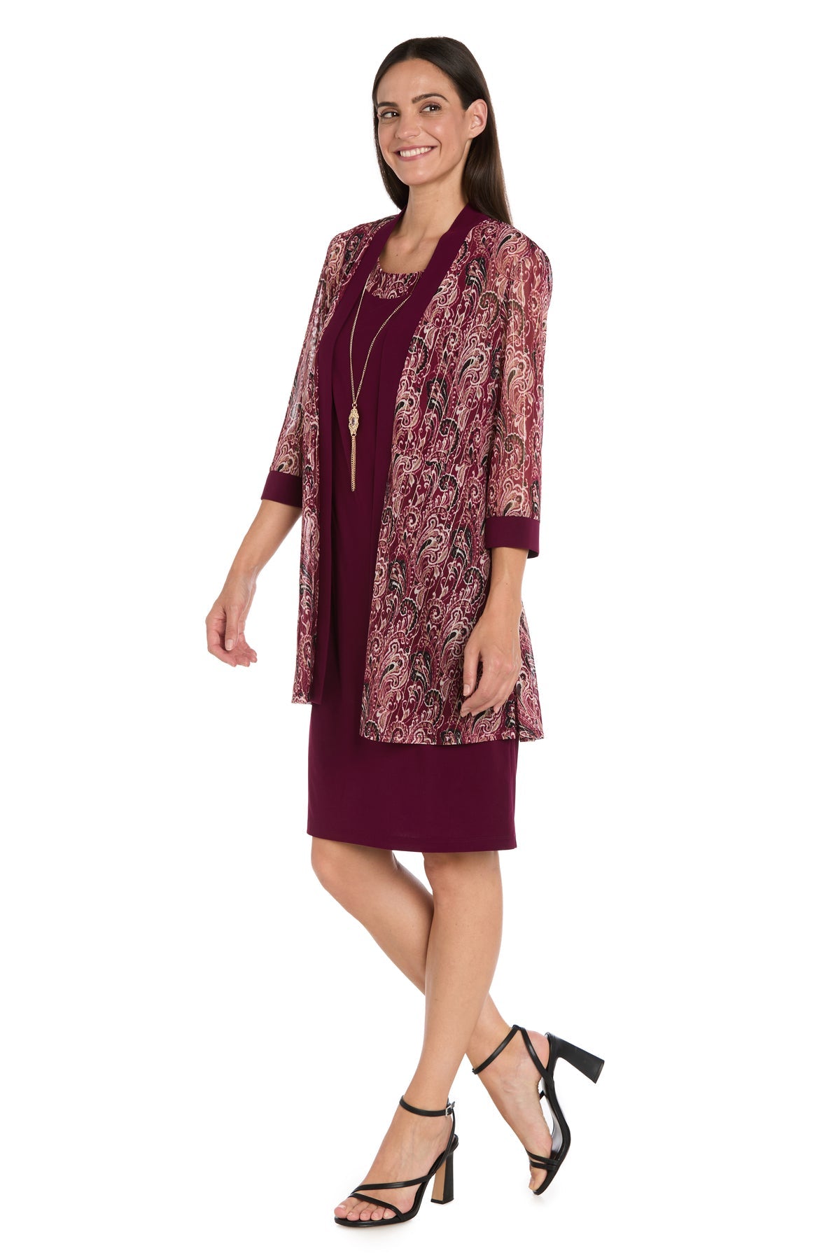 A woman stands against a white background wearing the Petite Short Wine Party Dress: R&M Richards 3029P, paired with a patterned sheer jacket, black heeled sandals, and a long necklace.